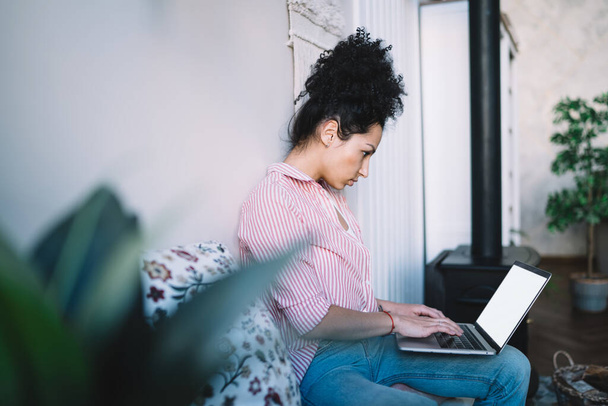 Side view of serious focused ethnic woman in trendy jeans using laptop while having rest cross legged on couch at home  - Foto, imagen