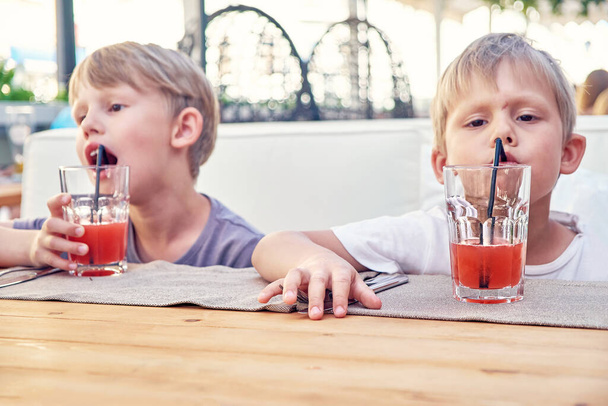 Cute little blond brothers drink fresh strawberry juice with straws sitting at wooden table in cafe in summer evening close view - Foto, imagen