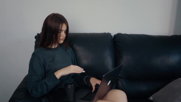 Деловая женщина работает дома онлайн сидя на диване, она выглядит заботливой и сосредоточенной на бизнесе. Одета в темно-зеленый чемодан, сидит на черном диване. Концепция работы. Концепция 4k - Кадры, видео