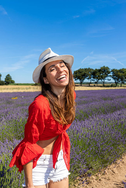 Muotokuva nuoresta naisesta leikkimässä ja nauramassa laventelipellolla. Hänellä on punainen pusero, valkoiset shortsit ja hattu.. - Valokuva, kuva