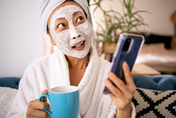 Szczęśliwy zaskoczony Azjatka zastosować maskę twarzy i za pomocą telefonu komórkowego dostał dobrą wiadomość, gdy siedzi na kanapie w domu. Pielęgnacja skóry i wellness poranna koncepcja. - Zdjęcie, obraz