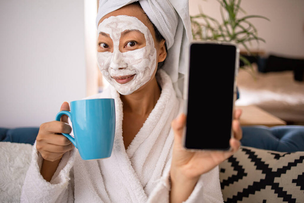 Boldog meglepett ázsiai nő alkalmazni arc maszk és mutatja okostelefon van egy jó hír, amikor ül a kanapén otthon. Szépség bőrápolás és wellness reggeli koncepció. - Fotó, kép