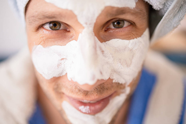 Hombre barbudo divertido con una máscara cosmética en su cara en albornoz y una toalla en su cabeza hace tratamientos de spa en su cara descansando en casa. - Foto, imagen