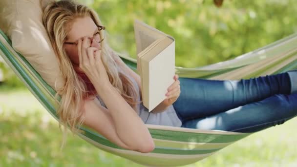 Lachende blonde vrouw met bril die een boek leest, ontspannen op de hangmat in de tuin, vrije tijd en zomervakantie concept - Video
