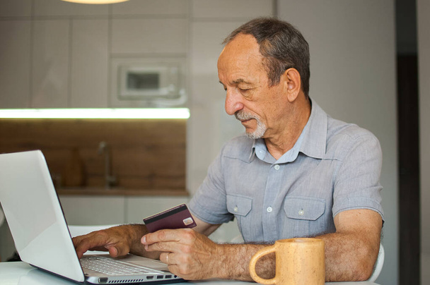 Trendy mature man is working from home with laptop sitting at the table in his kitchen, buying something with credit card online, happy retirement, home-office concepts - Foto, imagen