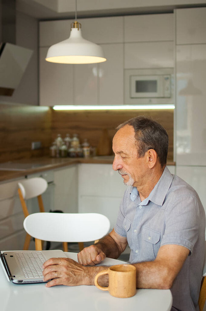 Trendy mature man is working from home with laptop sitting at the table in his kitchen, happy retirement, home-office concepts - Photo, Image