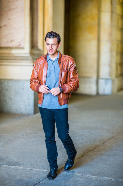 Dressing in a brown leather jacket, gray undershirt, black pants, one hand holding a cell phone,  a young handsome guy is walking and passing by a business building. - Photo, Image