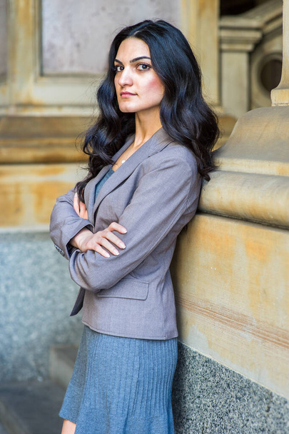 Dressing in a gray knit under dress and a jacket, crossing arms, a young businesswoman is standing outside an office building, looking sideways, relaxing and thinkin - Foto, afbeelding