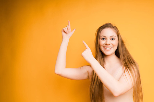 Egy vonzó lány, hosszú hajjal széles körben mosolyog, és az ujjaival jobbra mutat, elszigetelve a narancssárga háttértől. - Fotó, kép