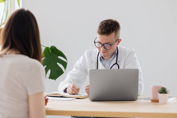 a young adult European and competent doctor in a white coat in a medical clinic in his office is seeing a patient - Photo, image