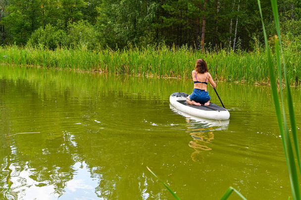 女性が密集した草に囲まれた狭い運河を通ってスープボード上を走行します。アクティブな週末の休暇野生の自然屋外。女性はジーンズで膝の上に座っている。夏. - 写真・画像