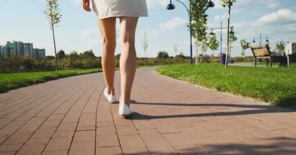 Kadın bacakları, arka görüş, gün boyunca parkta yürümek, batan güneşin ışınları bacaklara düşer, bir gölge oluşturur, yavaşlar, orta büyüklükte görüntüler - Video, Çekim