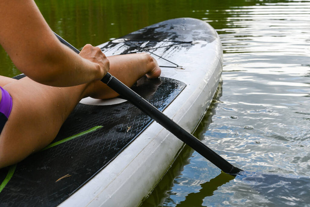 Eine Frau fährt auf dem Sup Board durch einen schmalen Kanal, der von dichtem Gras umgeben ist. Aktive Wochenendurlaube in freier Natur. Eine Frau sitzt mit ausgestreckten Beinen im Badeanzug. - Foto, Bild