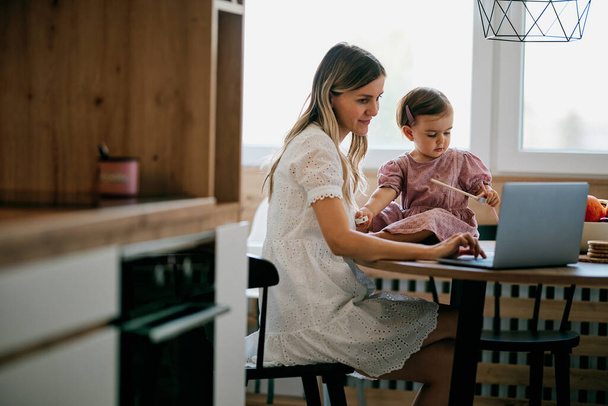 Bleiben Sie zu Hause in der Küche sitzen und arbeiten, während ihr kleines Mädchen ihr hilft. - Foto, Bild