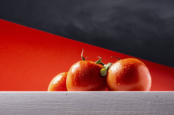 Lähikuva klusterin kolme märkää punaista luonnollista tomaattia valkoisella pöydällä punaisella taustalla ja osa taivasta. Valokuvassa on kopiotila ja se on otettu vaakasuorassa muodossa. - Valokuva, kuva