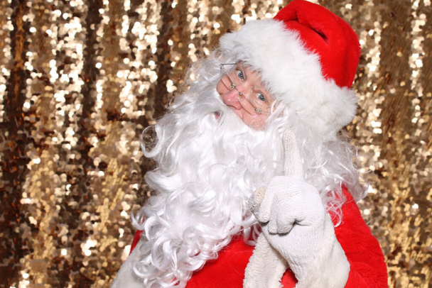 Santa Claus. Santa Claus holds his Finger in the air as he says DO NOT FORGET TO LEAVE MILK AND COOKIES FOR ME ON CHRISTMAS EVE. Gold Sequin background. Christmas Holiday Images. - Foto, Imagen