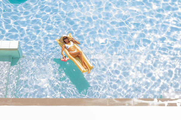 Vista superior de la chica en un traje de baño y sombrero flotando en el colchón de oro inflable en la piscina con cóctel de bebida. Vacaciones de verano y concepto de relax - Foto, imagen