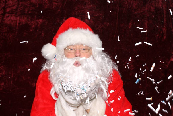 Santa Claus. Santa Claus blows shredded paper from his hands in a photo booth with a red velvet backdrop - Φωτογραφία, εικόνα