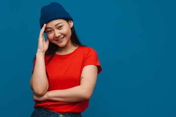 Asian teen girl in hat smiling and looking at camera isolated over blue background - Fotó, kép