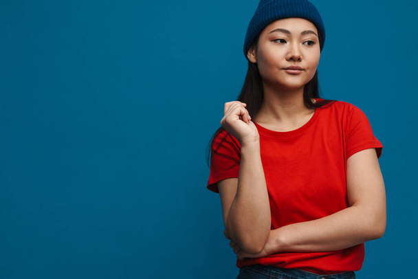 Asian teen girl in hat posing and looking aside isolated over blue background - Fotografie, Obrázek