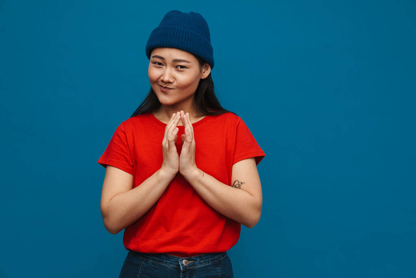 Asian teen girl in hat holding palms together and smiling isolated over blue background - Фото, изображение