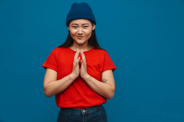 Asian teen girl in hat holding palms together and smiling isolated over blue background - Φωτογραφία, εικόνα