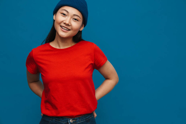 Asian teen girl in hat smiling and looking at camera isolated over blue background - Photo, image