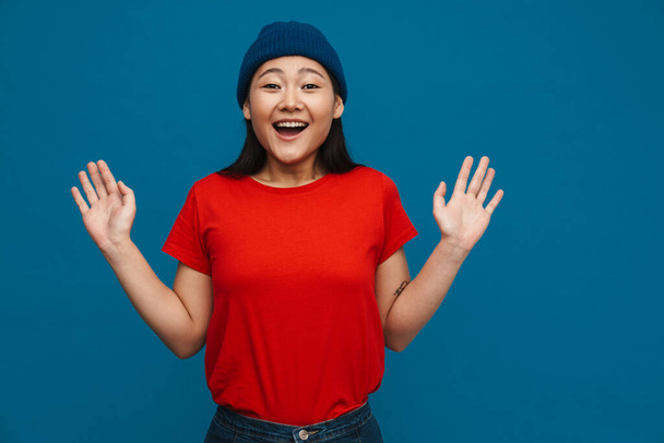 Excited asian teen girl in hat gesturing and smiling at camera isolated over blue background - 写真・画像