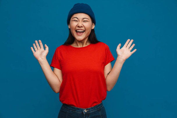 Excited asian teen girl in hat gesturing and laughing at camera isolated over blue background - Foto, Imagem