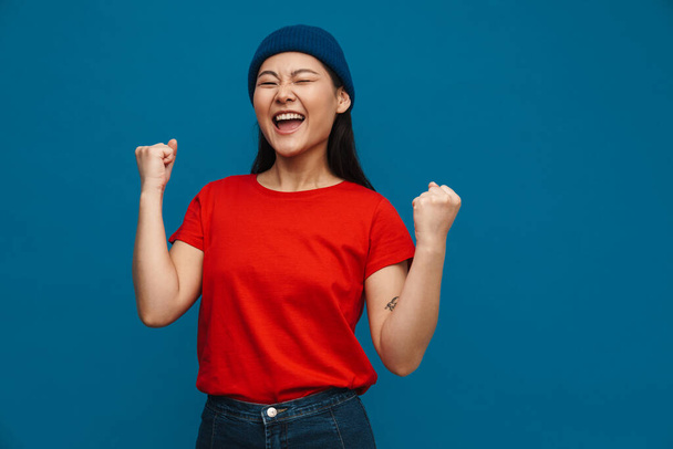 Excited asian teen girl in hat making winner gesture and screaming isolated over blue background - Foto, immagini