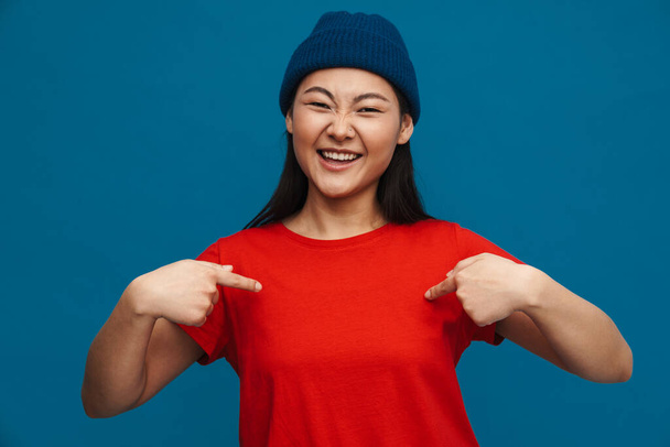 Asian teen girl in hat smiling and pointing fingers at herself isolated over blue background - Foto, Imagen