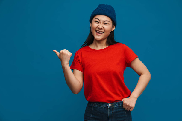 Happy young asian woman in casual clothes standing over blue wall background, pointing finger aside at copy space - Valokuva, kuva
