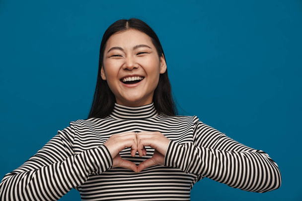 Laughing asian teen girl showing heart gesture at camera isolated over blue background - Foto, Imagem