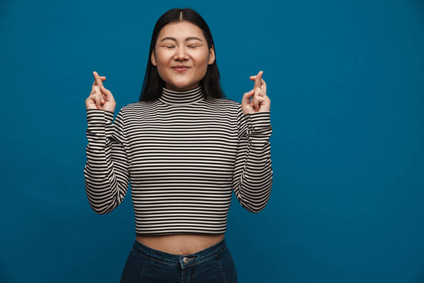 Smiling asian teen girl holding fingers crossed for good luck isolated over blue background - Foto, Imagen