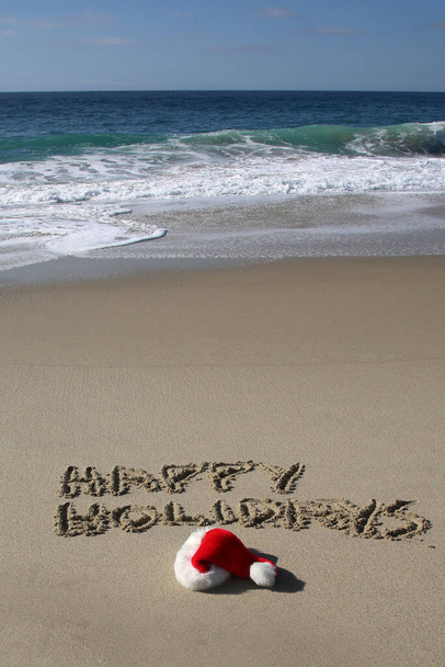 Santa Hat. Santa Claus hat on the beach with the words HAPPY HOLIDAYS written in the sand. Room for text. Words can be removed and replaced with your own. - Foto, afbeelding