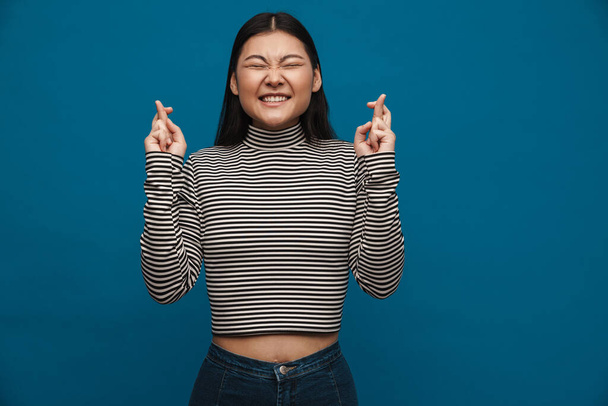 Smiling asian teen girl holding fingers crossed for good luck isolated over blue background - Foto, immagini