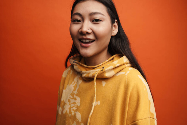 Asiática adolescente chica usando sudadera con capucha sonriendo y mirando a la cámara aislada sobre fondo naranja - Foto, imagen