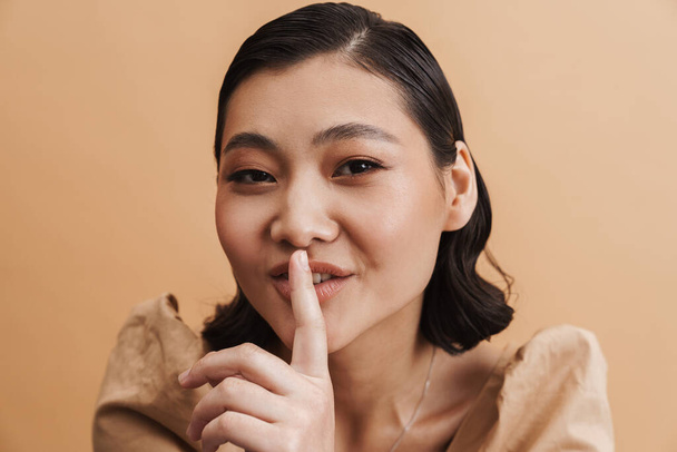 Young asian brunette woman showing silence gesture at camera isolated over beige background - Photo, image