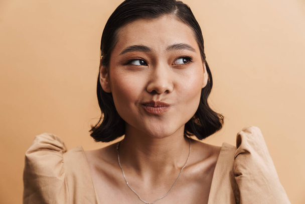 Young asian brunette woman grimacing and looking aside isolated over beige background - Foto, imagen