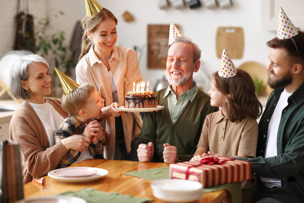 Kafkasyalı mutlu bir aile parti şapkası takıyor. Büyükbabalarının doğum gününü içeride kutluyorlar. Büyükbaba elinde mumlarla pasta tutuyor. - Fotoğraf, Görsel