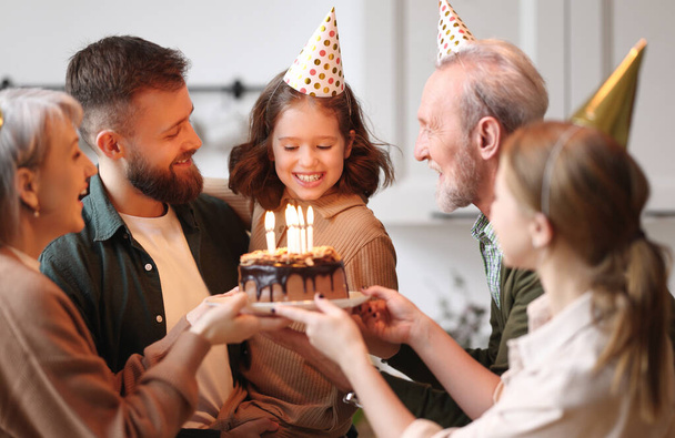かわいいです興奮した小さな女の子作る願いとケーキの上にキャンドルを吹いている間お祝いの誕生日で大きな美しい家族,正の両親と祖父母でパーティー帽子お祝いの幸せな女の子自宅 - 写真・画像