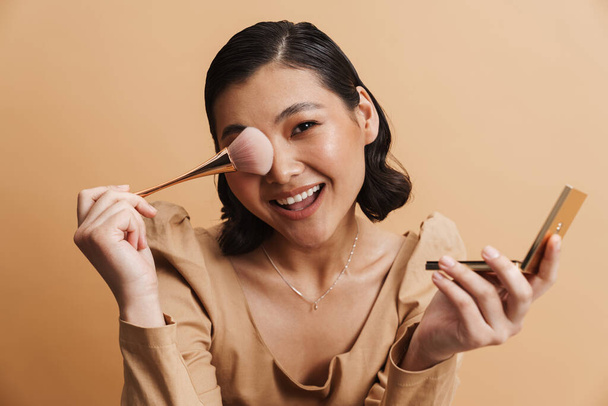 Smiling young brunette asian woman in dress posing standing holding makeup mirror over beige background holding blush brush - Foto, Imagen