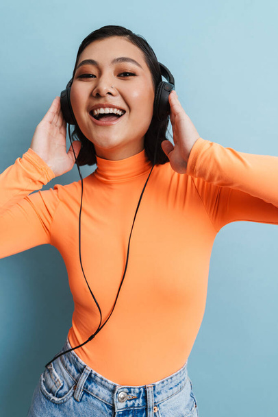feliz sorrindo jovem asiático morena mulher de pé sobre azul fundo ouvindo música com fones de ouvido - Foto, Imagem