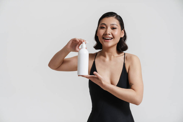 Brunette asian woman wearing swimsuit smiling and showing beauty product isolated over white background - Foto, immagini