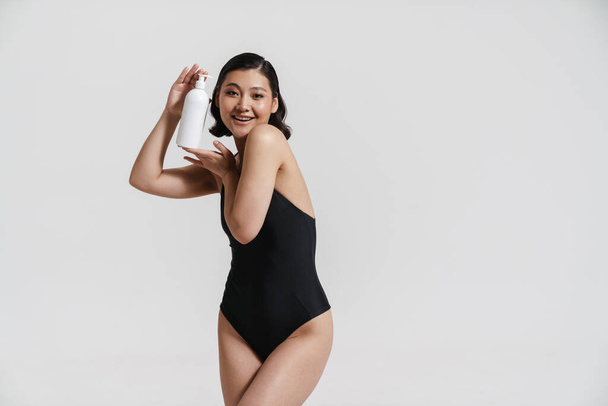Brunette asian woman wearing swimsuit smiling and showing beauty product isolated over white background - Foto, afbeelding