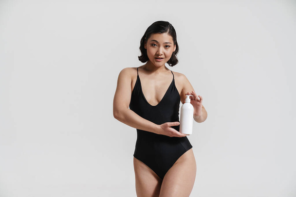 Brunette asian woman wearing swimsuit posing with beauty product isolated over white background - Fotó, kép