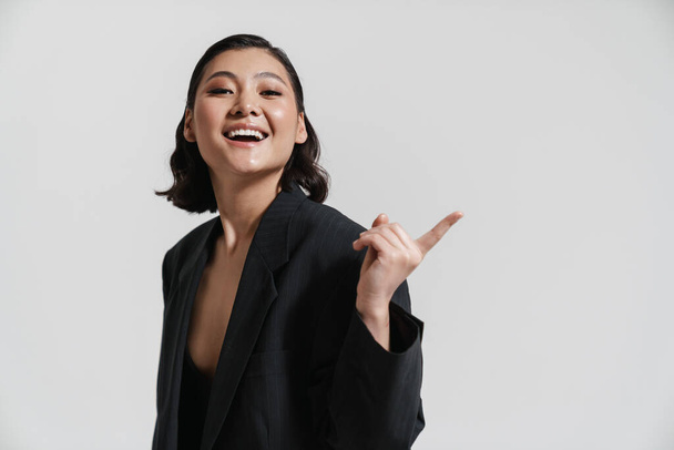 Smiling young asian woman wearing jacket standing over gray background pointing finger aside at copy space - Foto, immagini