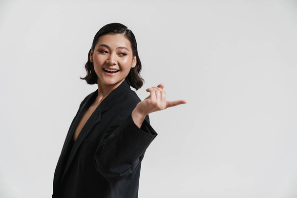 Sorridente giovane donna asiatica indossa giacca in piedi su sfondo grigio indicando dito da parte a copia spazio - Foto, immagini