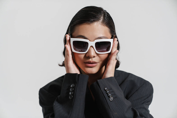 Young white asian woman in jacket posing with sunglasses over white wall background - Фото, изображение