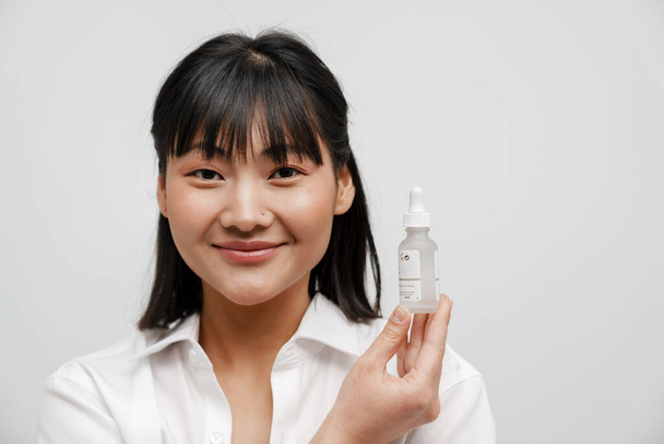 Young asian woman smiling while showing facial serum isolated over white background - Foto, immagini